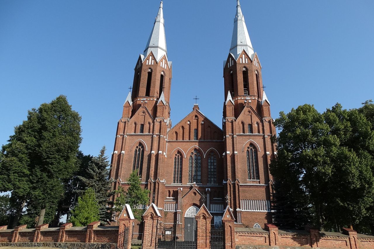 Raudonų plytų neogotikinė bažnyčia su dviem aukštais bokštais, žemiau žali medžiai ir mėlynas dangus
