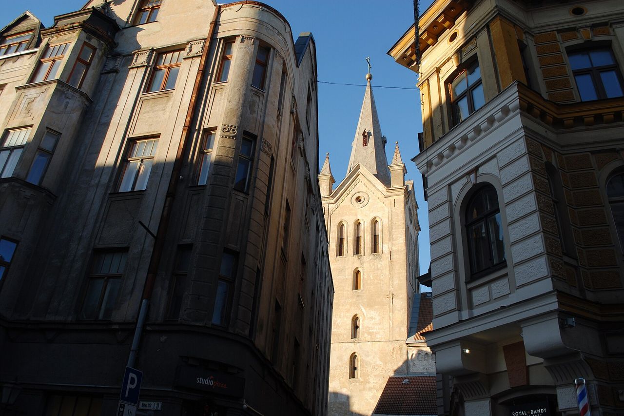 Šv. Jono bažnyčios bokštas su smaila viršūne ir apačioje siaura Rygos gatvė tarp senų pastatų Cėsyse