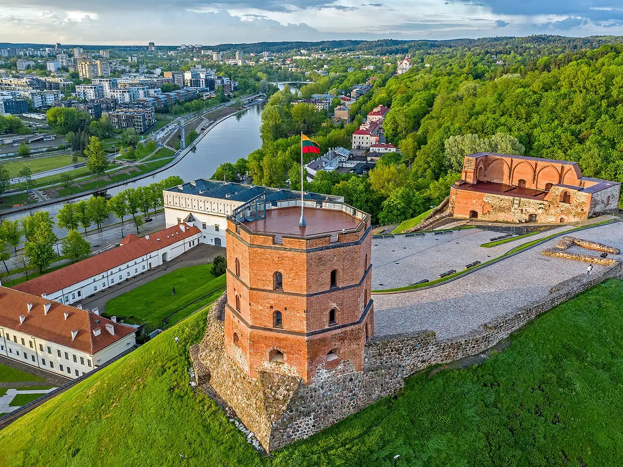 Raudonų plytų Gedimino pilies bokštas ant žaliuojančio kalno virš Neries, tolumoje miesto pastatai