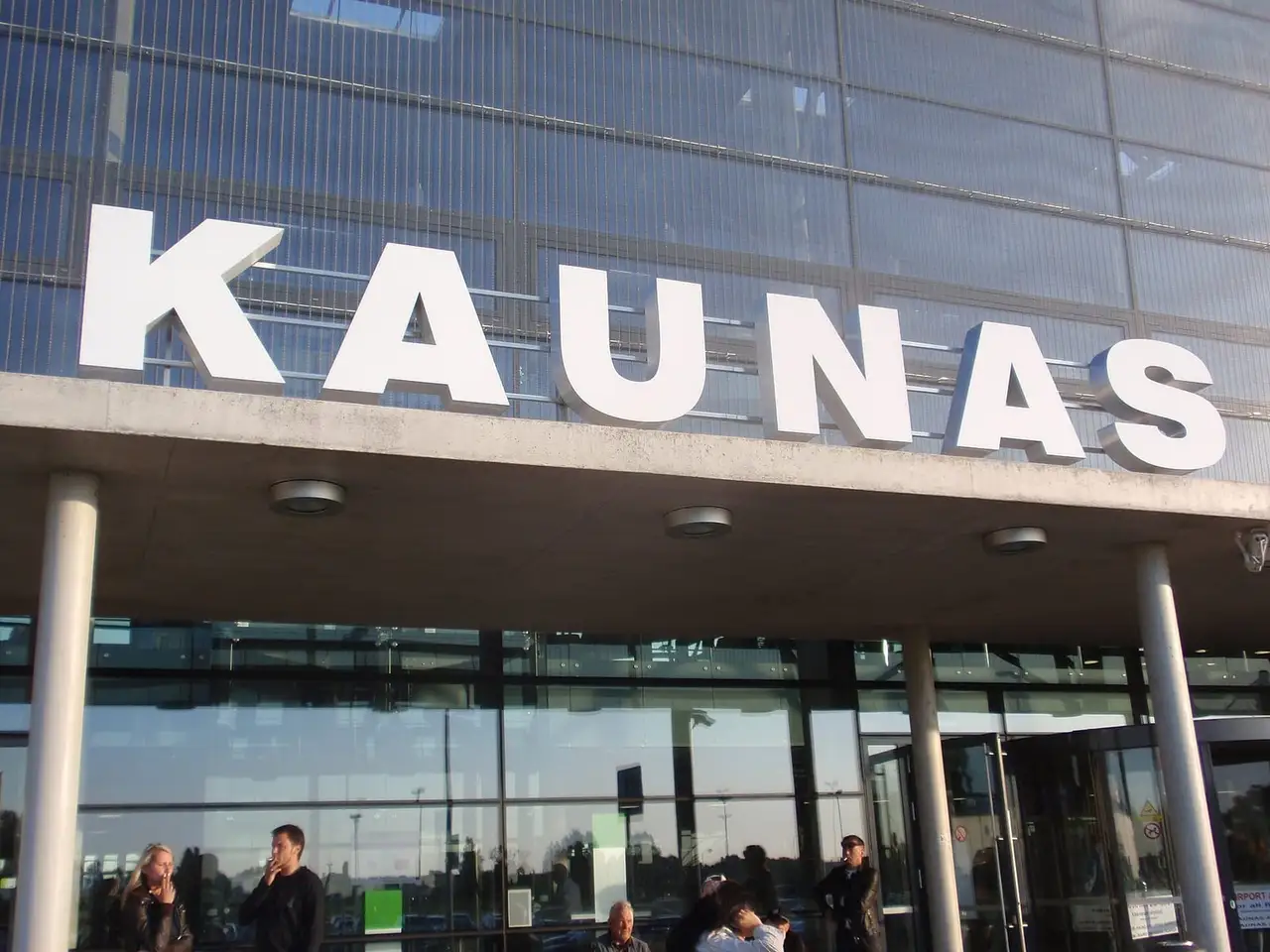 Baltos raidės „Kaunas“ ant oro uosto terminalo stogelio, po juo stikliniai įėjimo langai ir žmonės