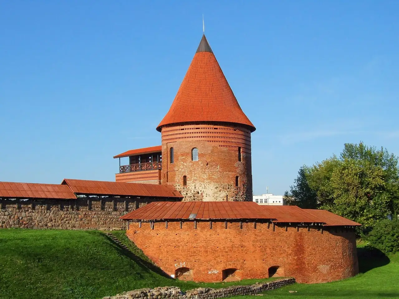 Raudonų plytų Kauno pilies bokštas su kūgiško stogu ir akmeninė siena prie žaliuojančio pylimo