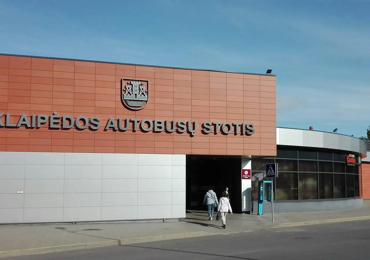 Klaipėdos autobusų stoties pastatas su miesto herbu ir užrašu virš įėjimo, priekyje praeina du keleiviai