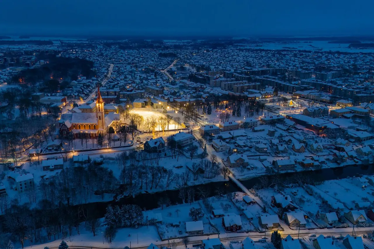 Žiemos vakaro Kretingos centras iš paukščio skrydžio, apšviesta bažnyčia ir sniegu padengti stogai