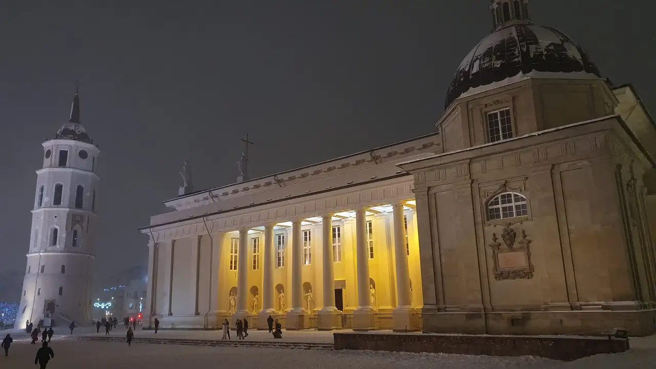 Sniegu padengta Vilniaus Katedros aikštė vakare, apšviesta arkikatedra su kolonomis ir varpinė