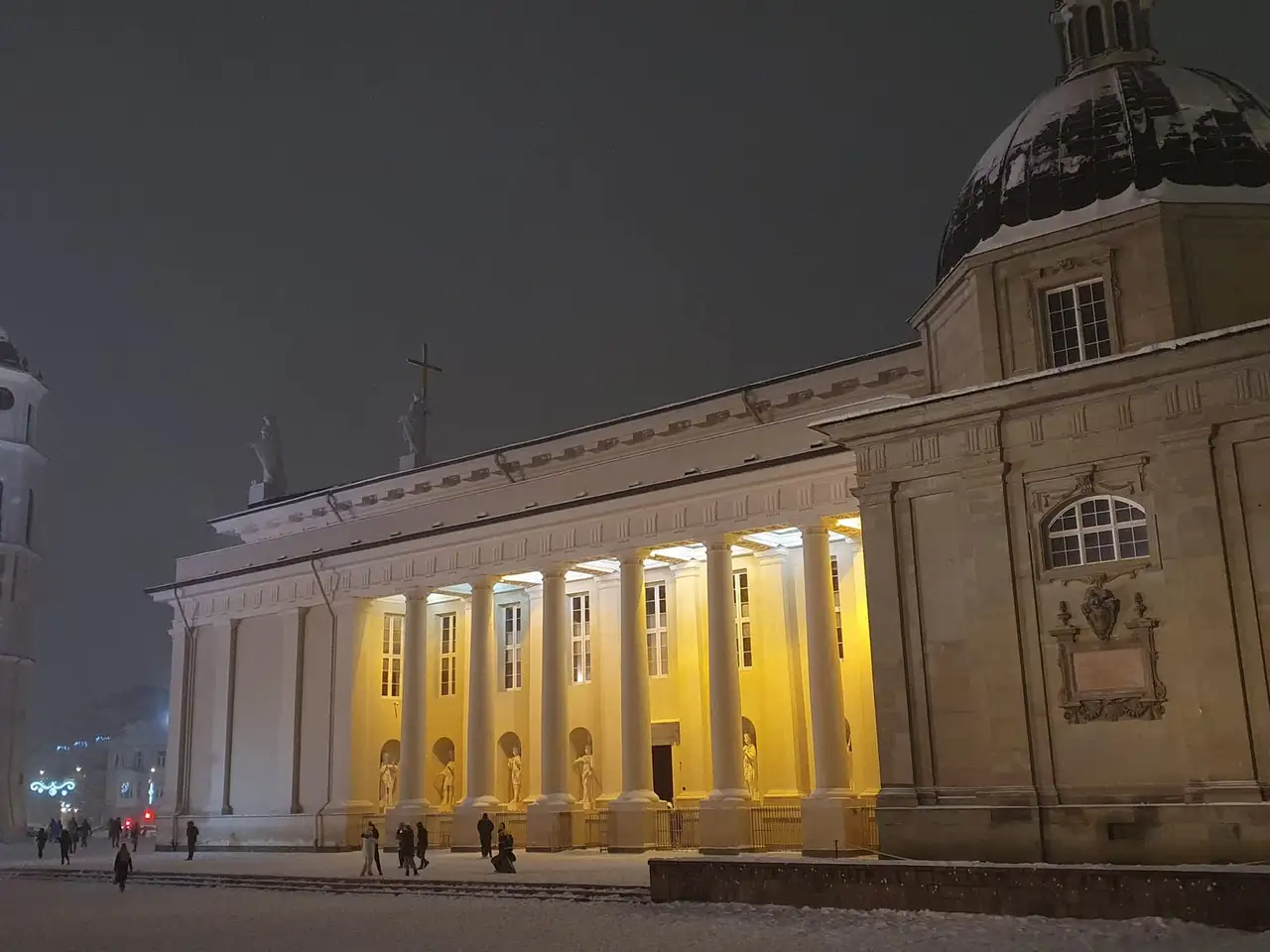 Sniegu padengta Vilniaus Katedros aikštė vakare, apšviesta arkikatedra su kolonomis ir varpinė