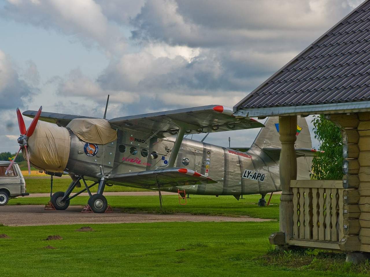 Sidabrinis lėktuvas An-2 su apdengtu sraigtu žolėje šalia rąstinio pastato