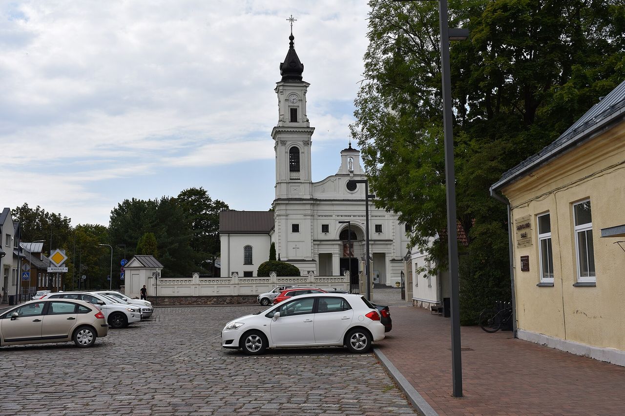 Balta Marijampolės bazilika su laikrodžio bokštu, priekyje akmenimis grįsta aikštė su automobiliais