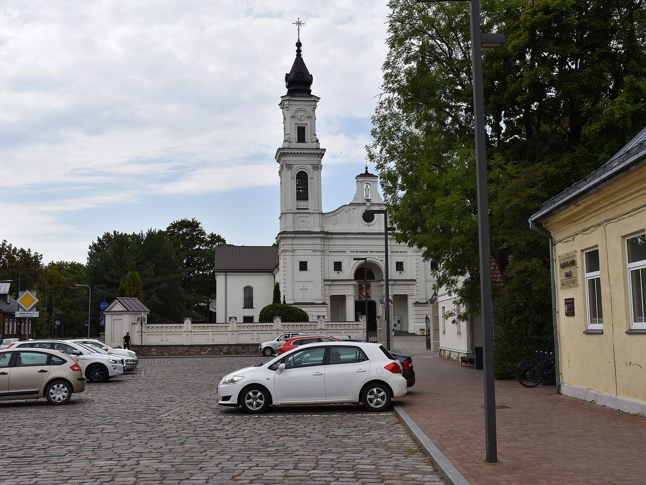 Balta Marijampolės bazilika su laikrodžio bokštu, priekyje akmenimis grįsta aikštė su automobiliais