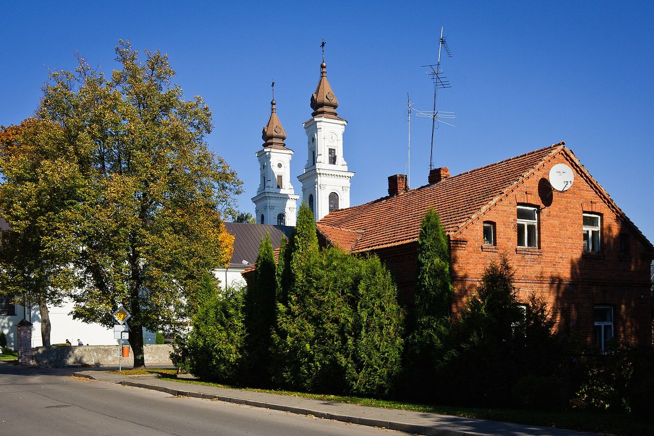 Marijampolės bazilikos balti bokštai virš raudonų plytų namo ir rudens medžių