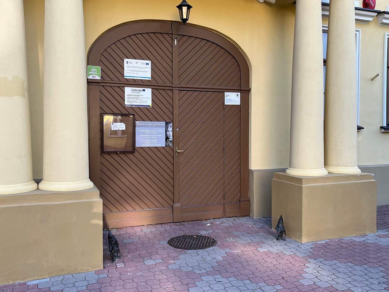 Rudos rąstinės durys su skliautu tarp geltonų kolonų, prie jų lentelė su muziejaus darbo laiku
