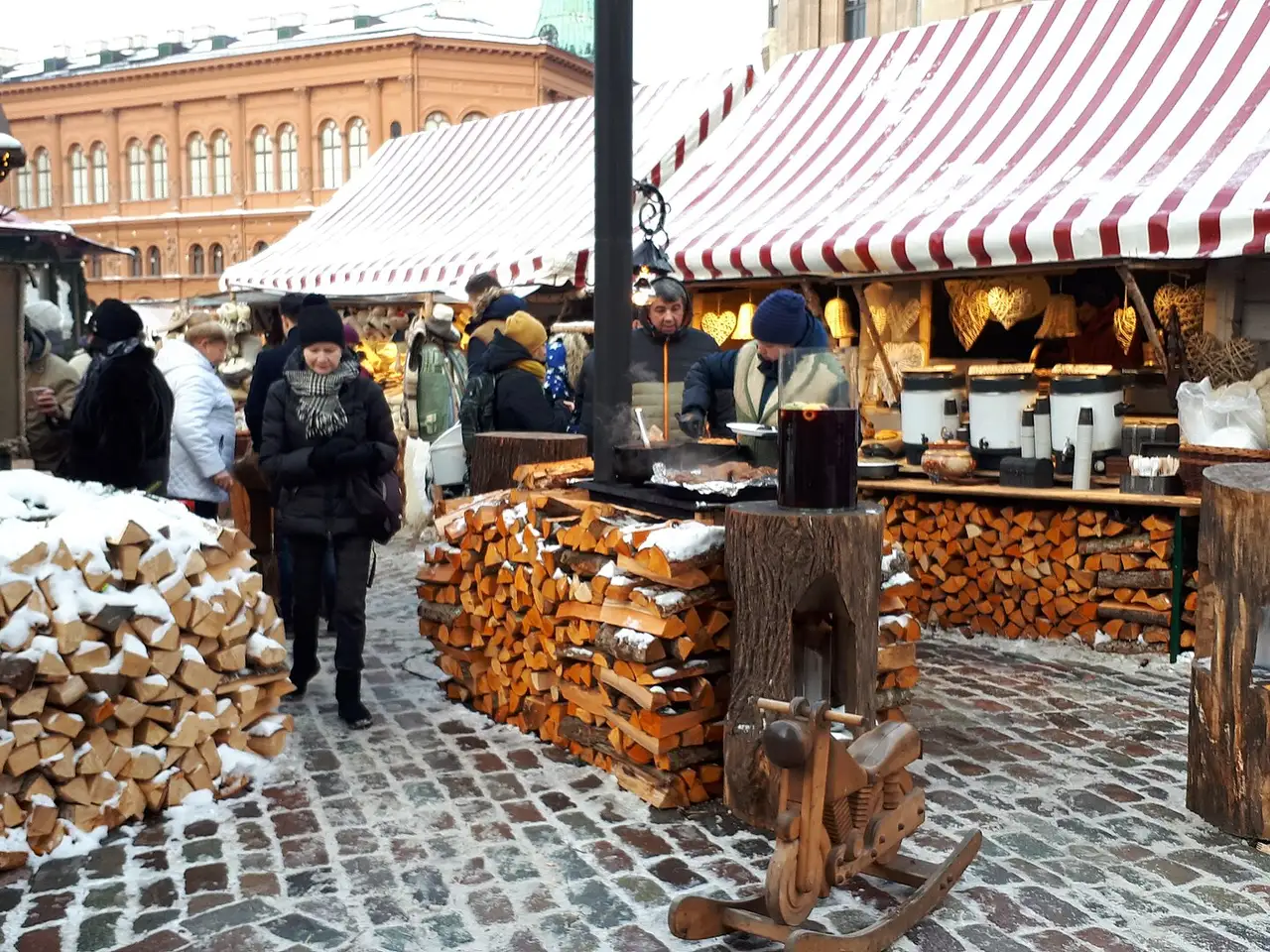 Šilto vyno kioskas Rygos Kalėdų turguje, sniegas ant grindinio ir sukrautos malkos