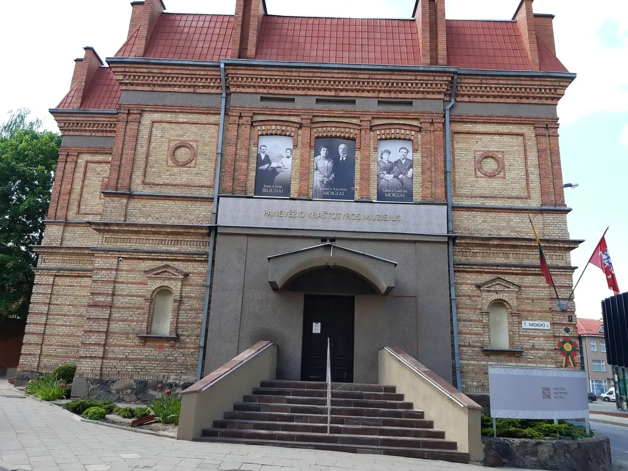 Raudonų plytų Panevėžio kraštotyros muziejaus fasadas su laiptais ir trimis nuotraukomis ant sienos