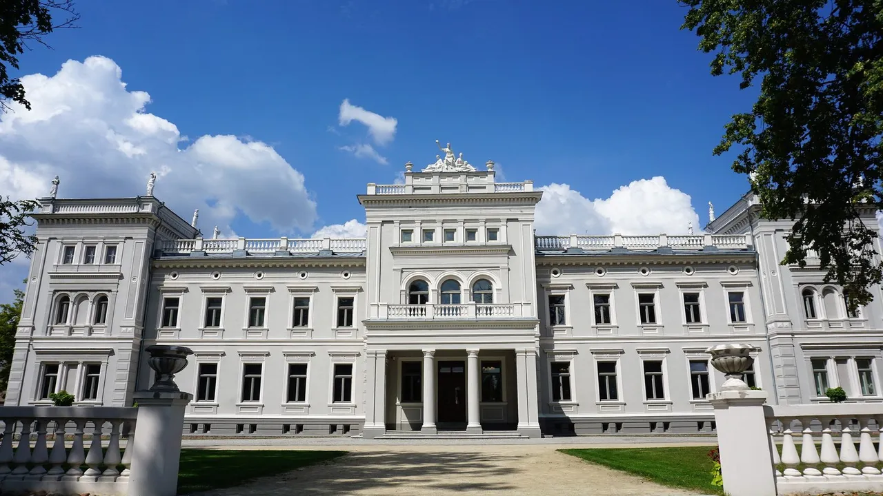Pilkšvi Plungės Oginskių rūmai su skulptūromis ant stogo ir akmeniniais laiptais prieš mėlyną dangų
