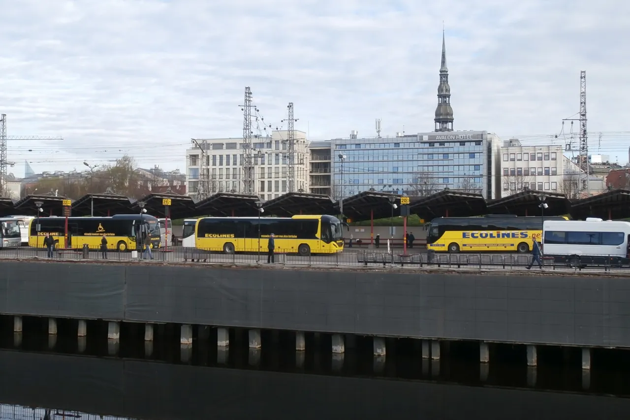 Geltoni tolimojo susisiekimo autobusai Rygos autobusų stotyje, už jų – Šv. Petro bažnyčios bokštas