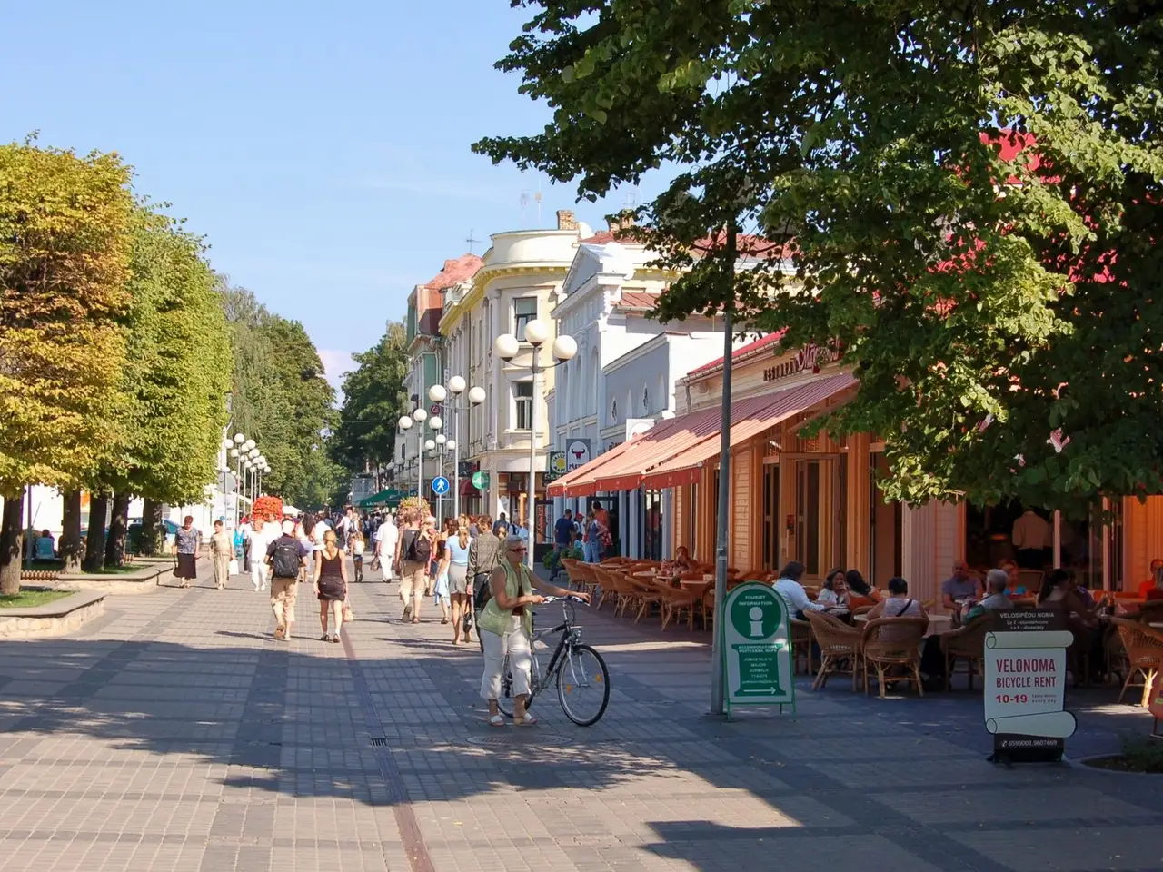 Pėsčiųjų Jomas gatvė Jūrmaloje su medžiais, kavinės terasa ir einančiais žmonėmis