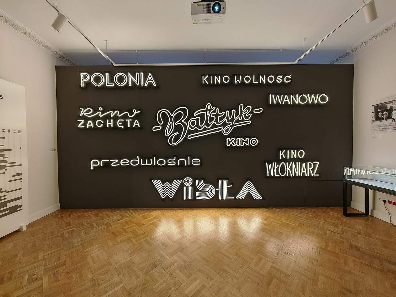 Šviečiančių neoninių kino pavadinimų siena Kinematografijos muziejaus salėje su parketo grindimis
