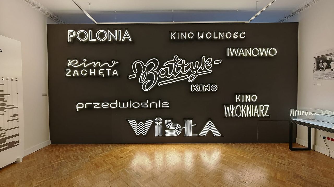 Šviečiančių neoninių kino pavadinimų siena Kinematografijos muziejaus salėje su parketo grindimis