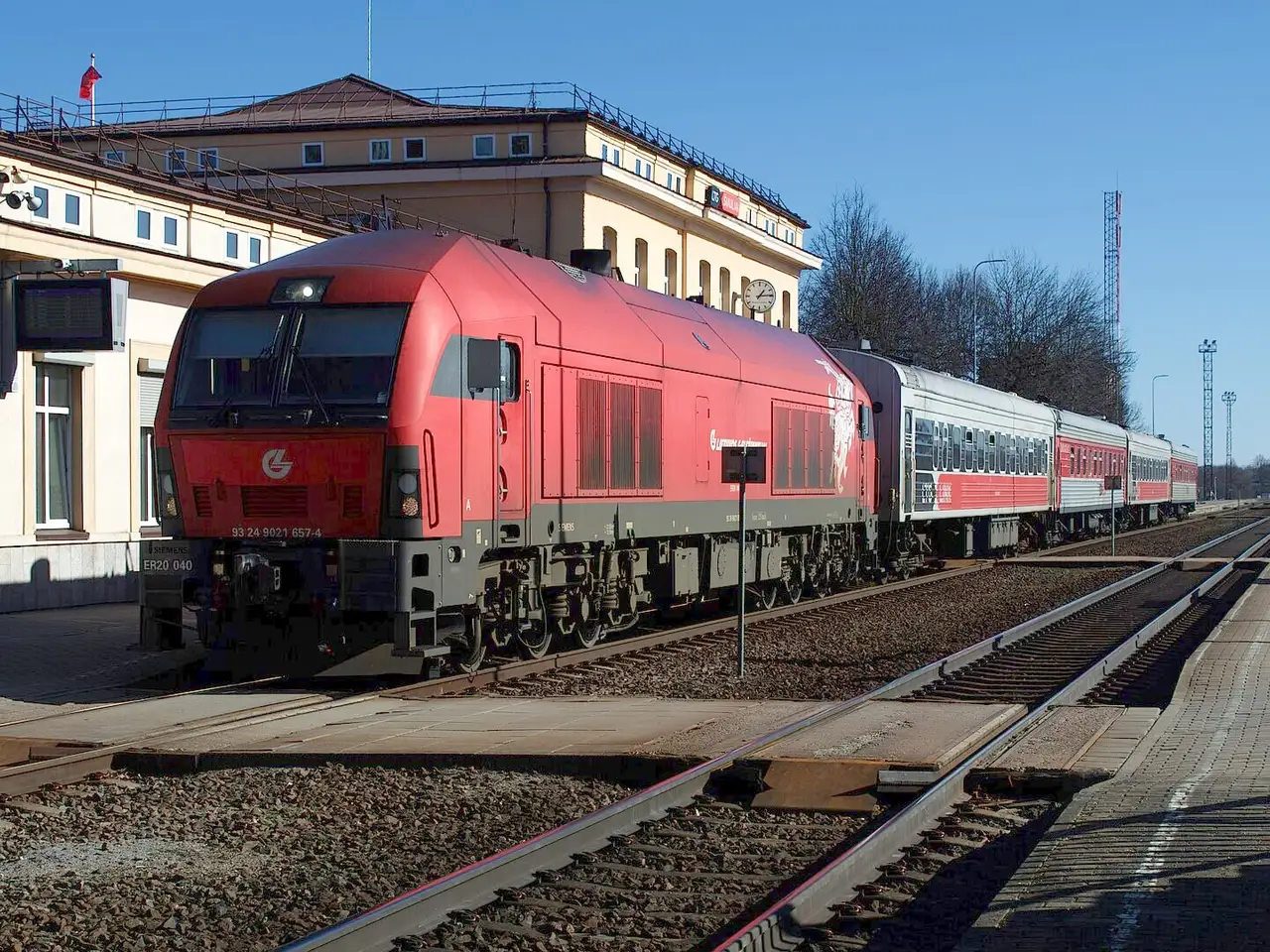 Raudonas lokomotyvas su keleiviniais vagonais Šiaulių geležinkelio stotyje giedrą dieną