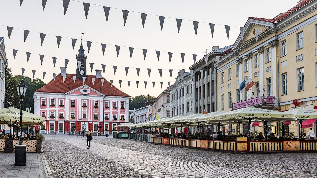 Tartu rotušės aikštė su rausvu rotušės pastatu, kavinių skėčiais ir iškilmingomis vėliavėlėmis