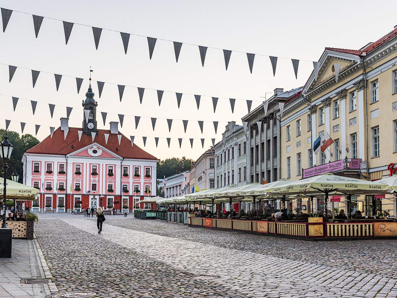Tartu rotušės aikštė su rausvu rotušės pastatu, kavinių skėčiais ir iškilmingomis vėliavėlėmis