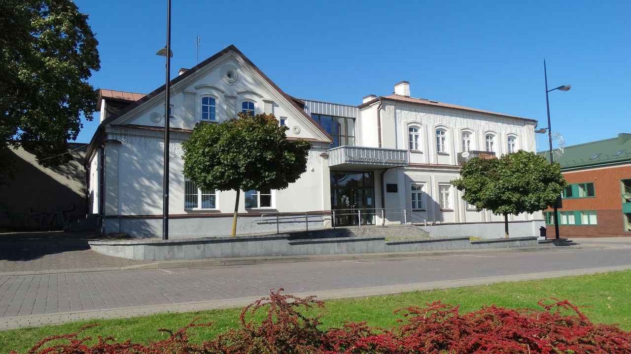 Šviesus Utenos kraštotyros muziejaus pastatas su dviem aukštais ir vešliais medžiais priekyje