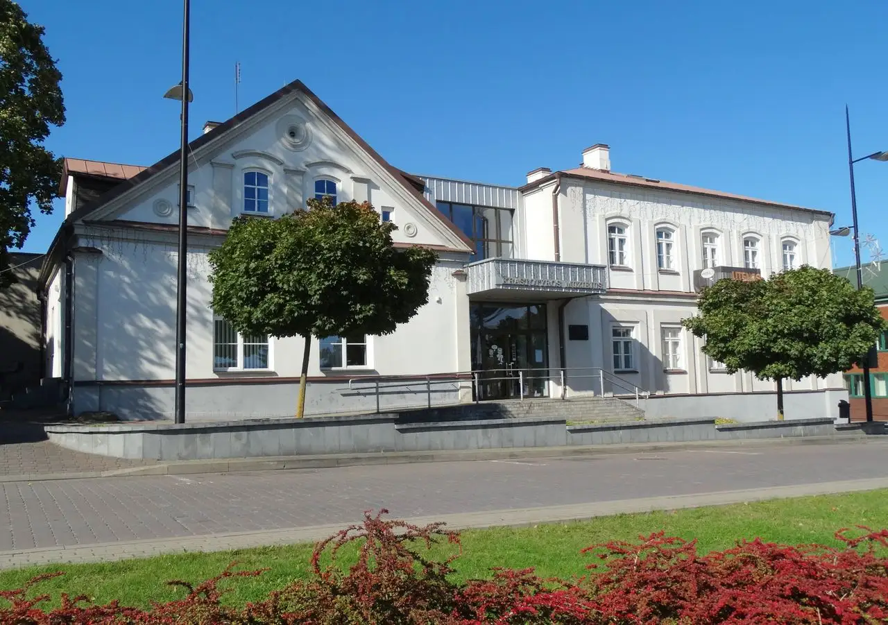 Baltas Utenos kraštotyros muziejaus pastatas su aukštu stogu, medžiais ir šaligatviu priekyje