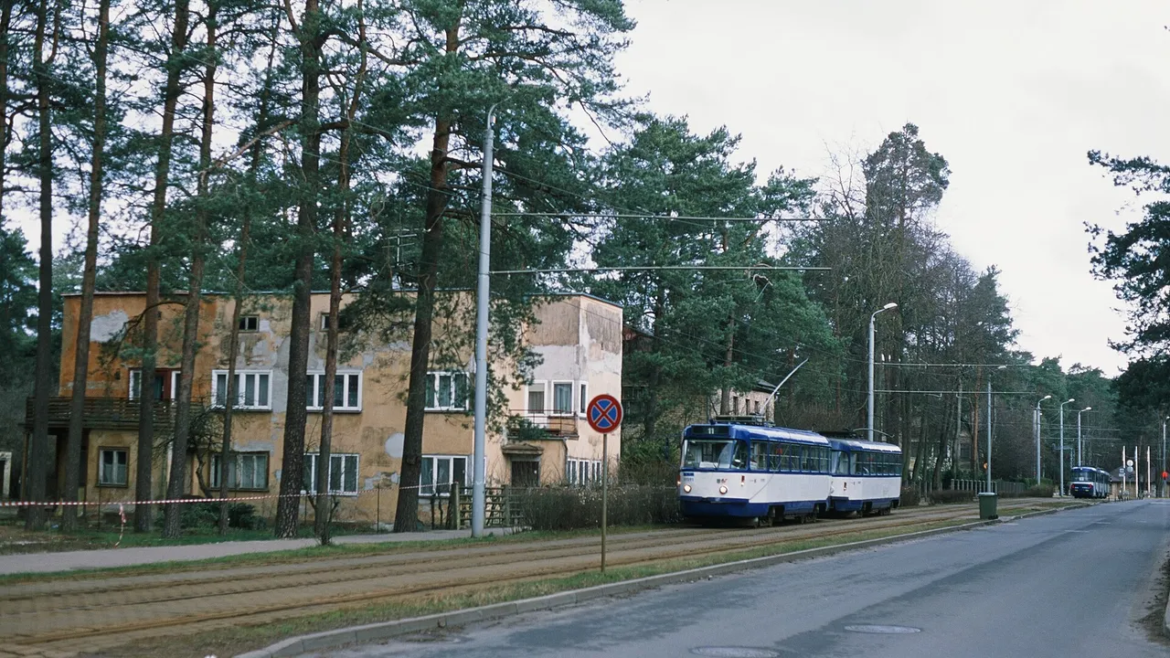 Mėlynai baltas Rygos tramvajus važiuoja gatve tarp pušų ir geltono namo