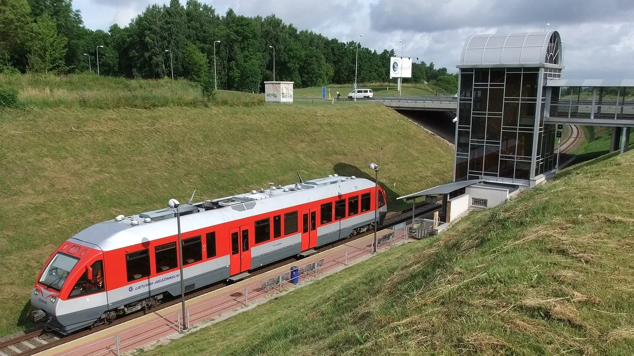 Raudonai pilkas dyzelinis traukinys oro uosto geležinkelio stotelėje, šalia stiklinis liftas ir žolėtas šlaitas