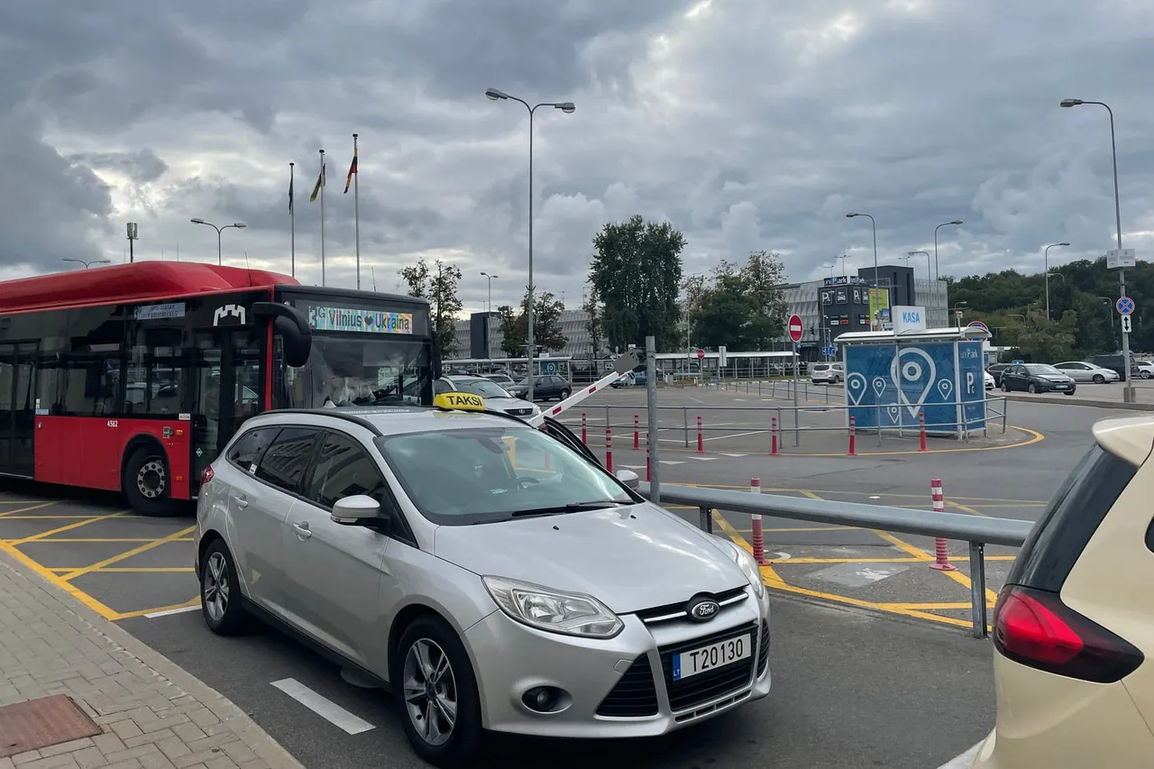 Raudonas miesto autobusas ir sidabrinis taksi automobilis aikštelėje prie Vilniaus oro uosto