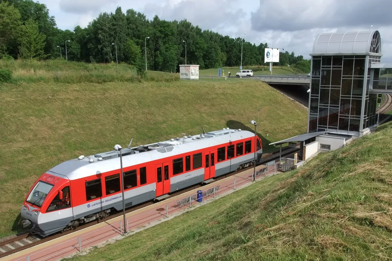 Raudonas dyzelinis traukinys oro uosto geležinkelio stotelėje, už jo matosi stiklinis liftas